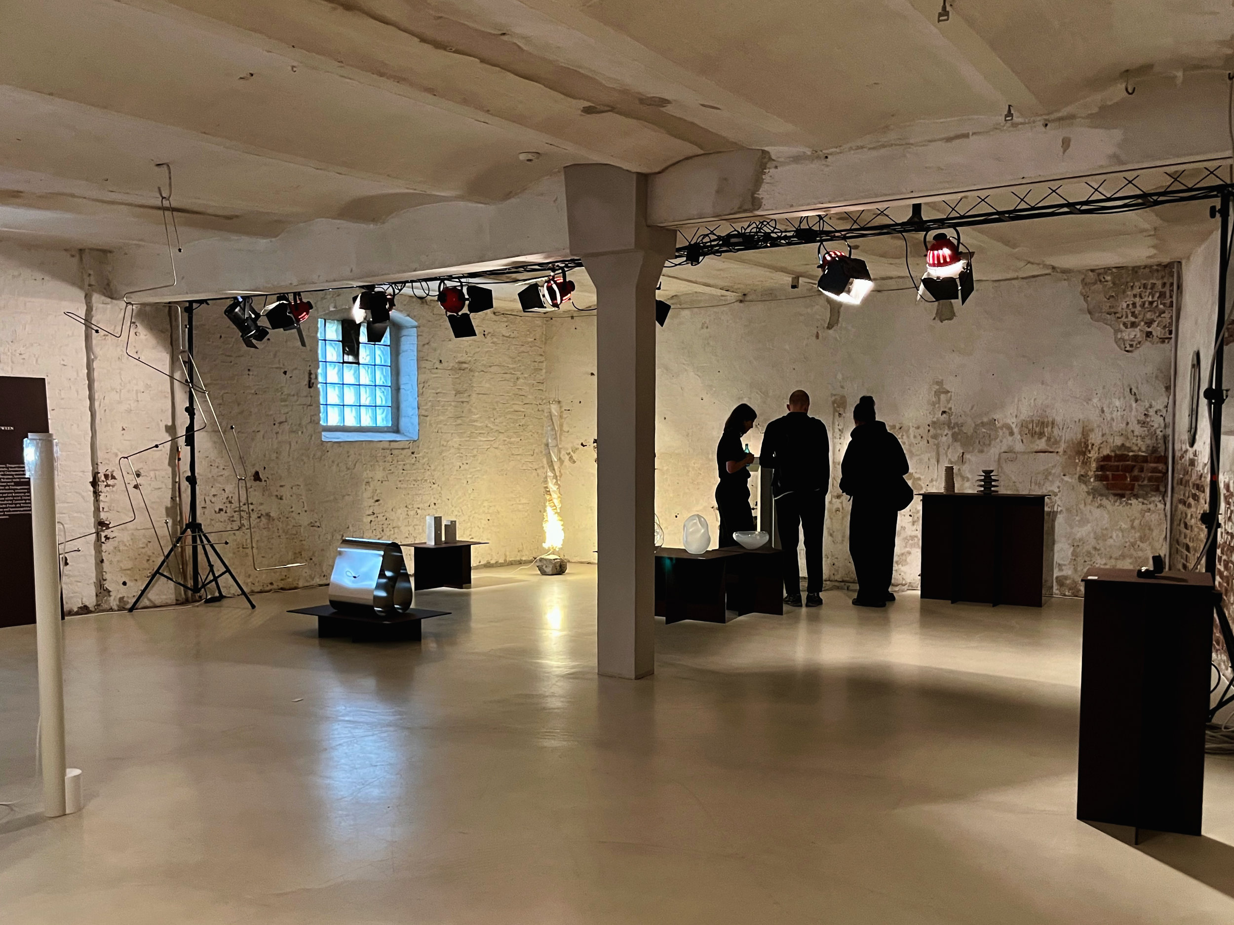 Eine Kunstausstellung. Die Ausstellungsfläche ist minimal eingerichtet. Es stehen vereinzelt Skulpturen im Raum. Drei Menschen schauen sich im Hintergrund eine Skulptur an.